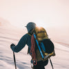 A skier viewed from behind on a snowy mountain slope, wearing a blue jacket and a large yellow and black backpack with a red rope and climbing gear attached. The sun is low on the horizon, creating a bright glow.