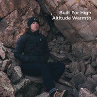 woman sits in rocky area wearing a black puffy down jacket.