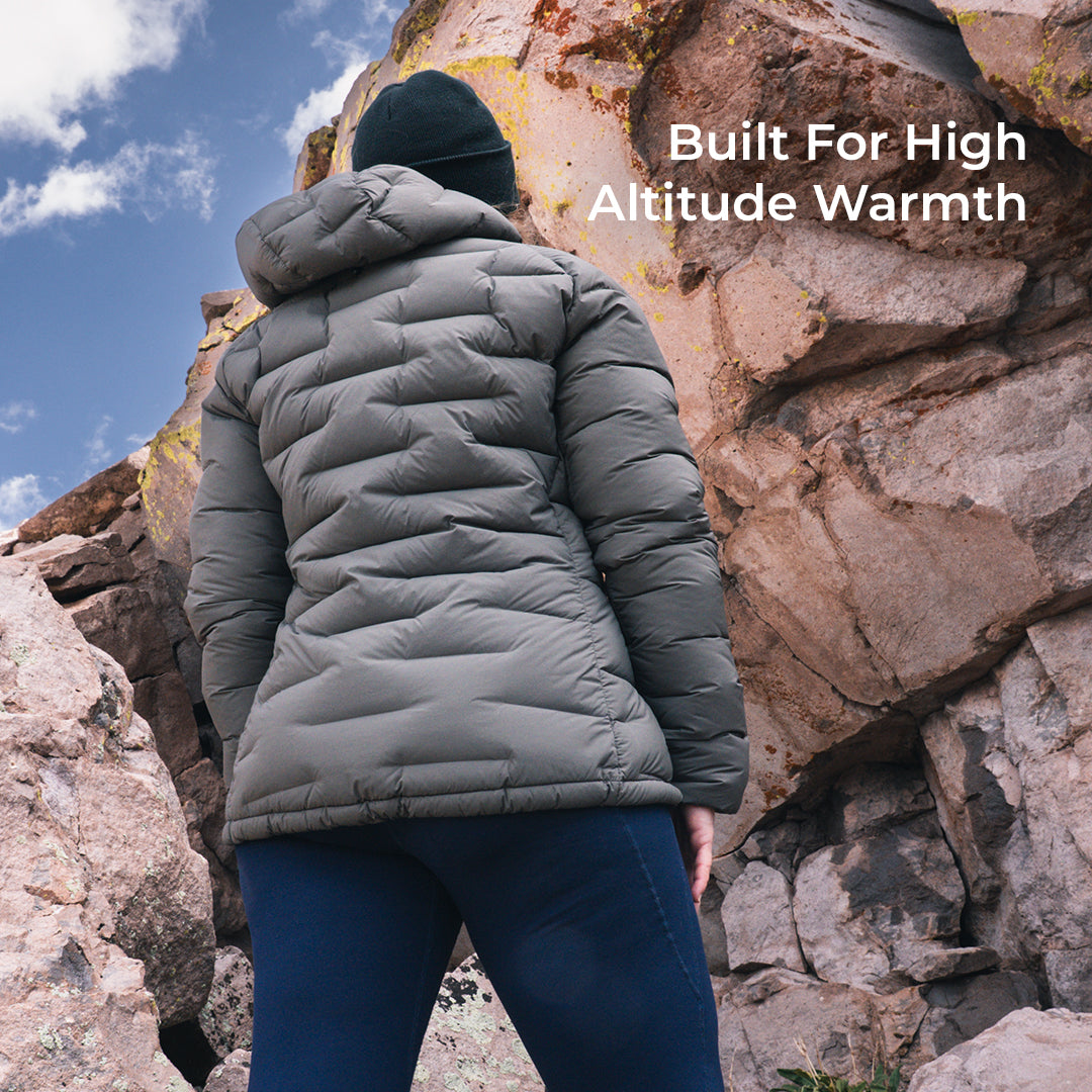 A person viewed from behind wearing a quilted puffer jacket in an outdoor rocky setting under a blue sky. White text on the right reads 'Built For High Altitude Warmth'.