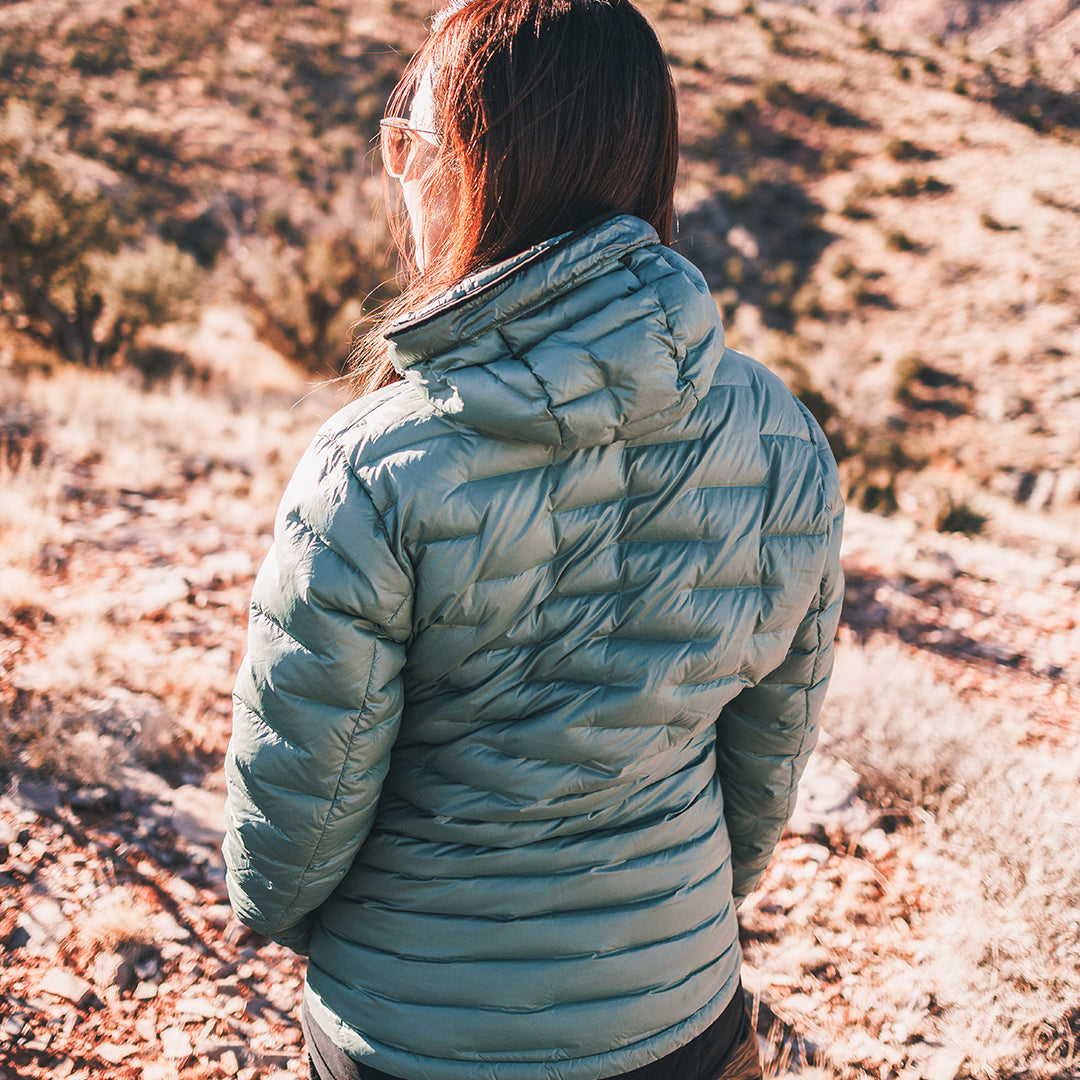 A person viewed from behind wearing a light sage green hooded puffer jacket, standing outdoors on a rocky, dry hillside.
