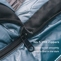 Close-up of a blue down sleeping bag zipper with black trim and a black pull tab, with white text reading 'No. 5 YKK Zippers sized to run smoothly easy to find in the dark.