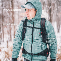 A man wearing a teal insulated winter jacket with black YKK zippers, shown outdoors in the snow with text callouts pointing to armpit vents and insulated pockets.