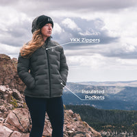 A woman wearing a dark gray puffer jacket stands on a rocky mountain overlook with a cloudy sky and forested hills in the background. White callout lines point to the jacket with text reading 'YKK Zippers' and 'Insulated Pockets.