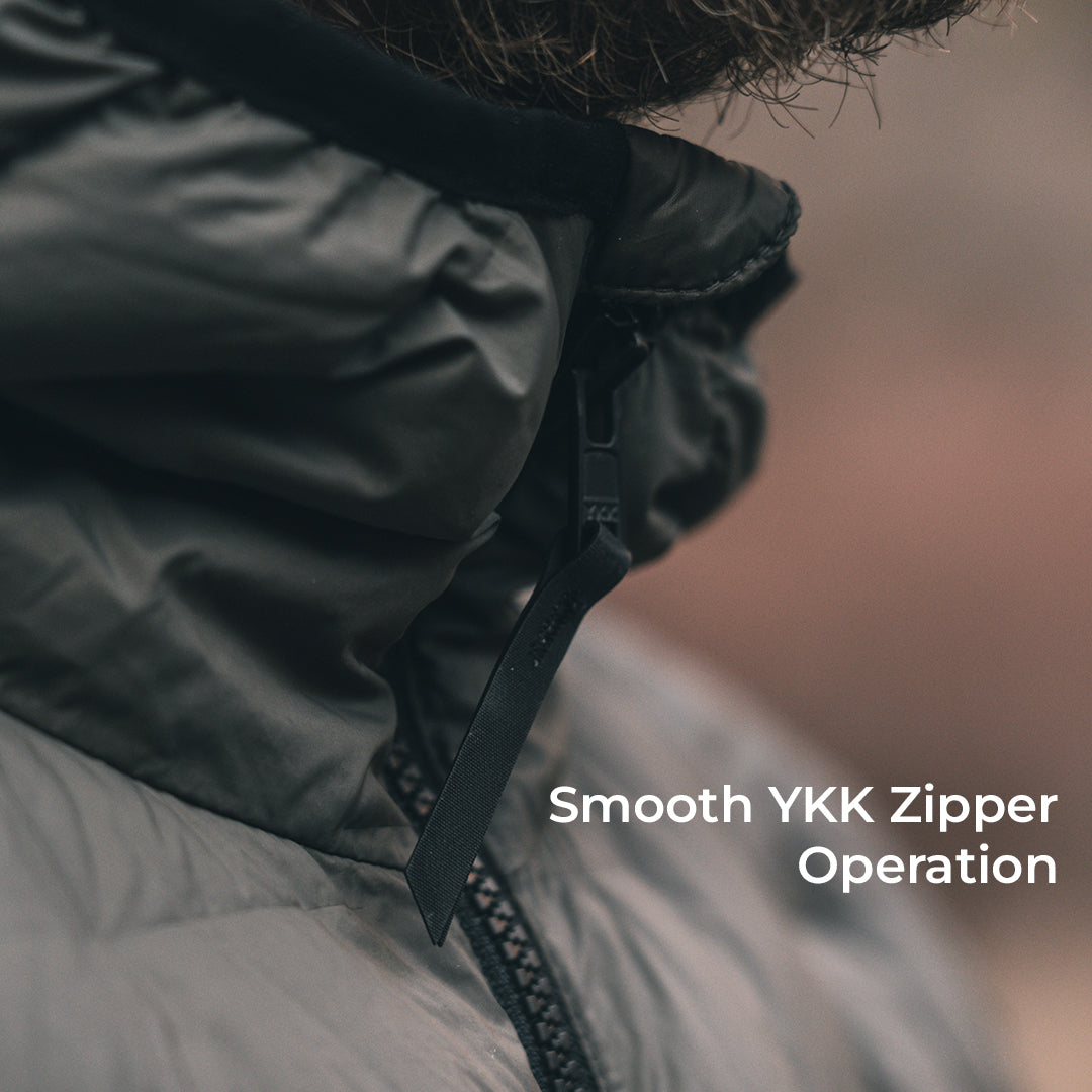 Close-up of a person wearing a dark gray puffer jacket, focusing on the zipper tab and pull tab, with the text 'Smooth YKK Zipper Operation' overlaid on the right side.