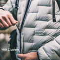 Close-up of a light gray puffer jacket with a black zipper being pulled, with text labels pointing to armpit vents, insulated pockets, and YKK zippers.
