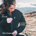 A woman outdoors on a rocky mountain setting wearing a black YKK jacket, demonstrating the full front zip and pointing to insulated pockets with text overlays.