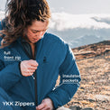 A woman outdoors in a mountain setting wearing a blue insulated jacket, with callout labels pointing to the front zipper and insulated pockets. Text at the bottom reads 'YKK Zippers'.