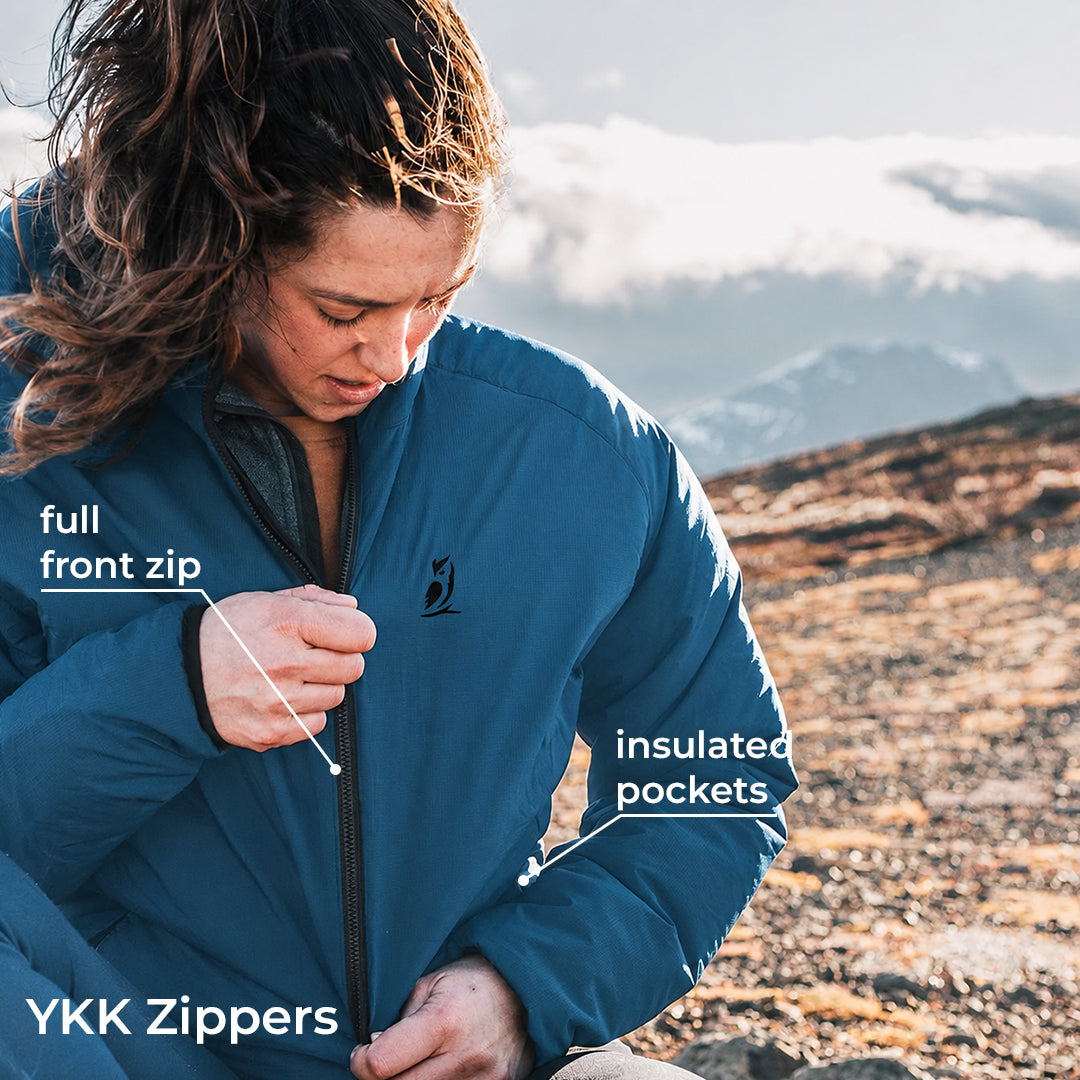 A woman outdoors in a mountain setting wearing a blue insulated jacket, with callout labels pointing to the front zipper and insulated pockets. Text at the bottom reads 'YKK Zippers'.