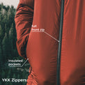 Close-up of a rust-orange insulated jacket with a full front zipper and labeled insulated pockets, shown outdoors with a blurred forest background. White text overlays point to the zipper and pockets.