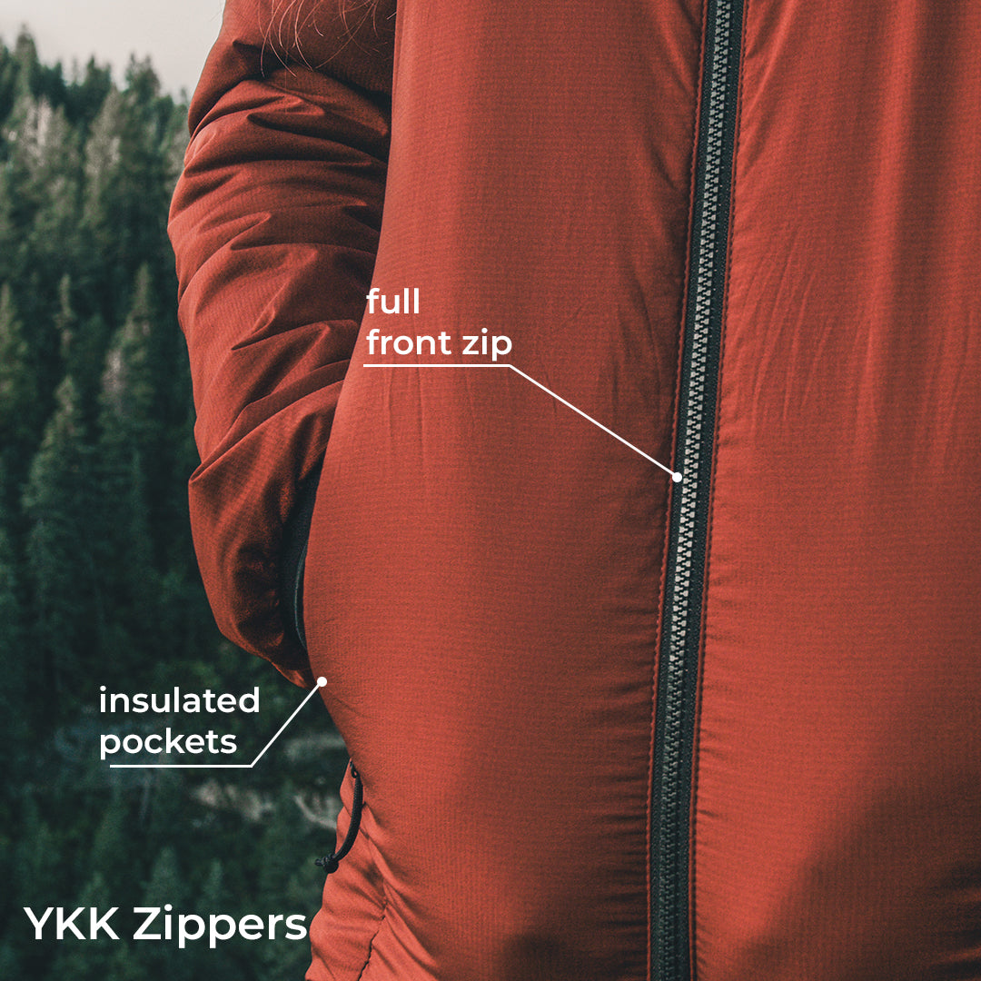 Close-up of a rust-orange insulated jacket with a full front zipper and labeled insulated pockets, shown outdoors with a blurred forest background. White text overlays point to the zipper and pockets.