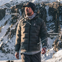 A man wearing a gray insulated puffer jacket stands in a snowy mountain landscape. White text labels point to the jacket, reading 'YKK Zippers' near the chest zipper area and 'Insulated Pockets' near the lower pocket area.