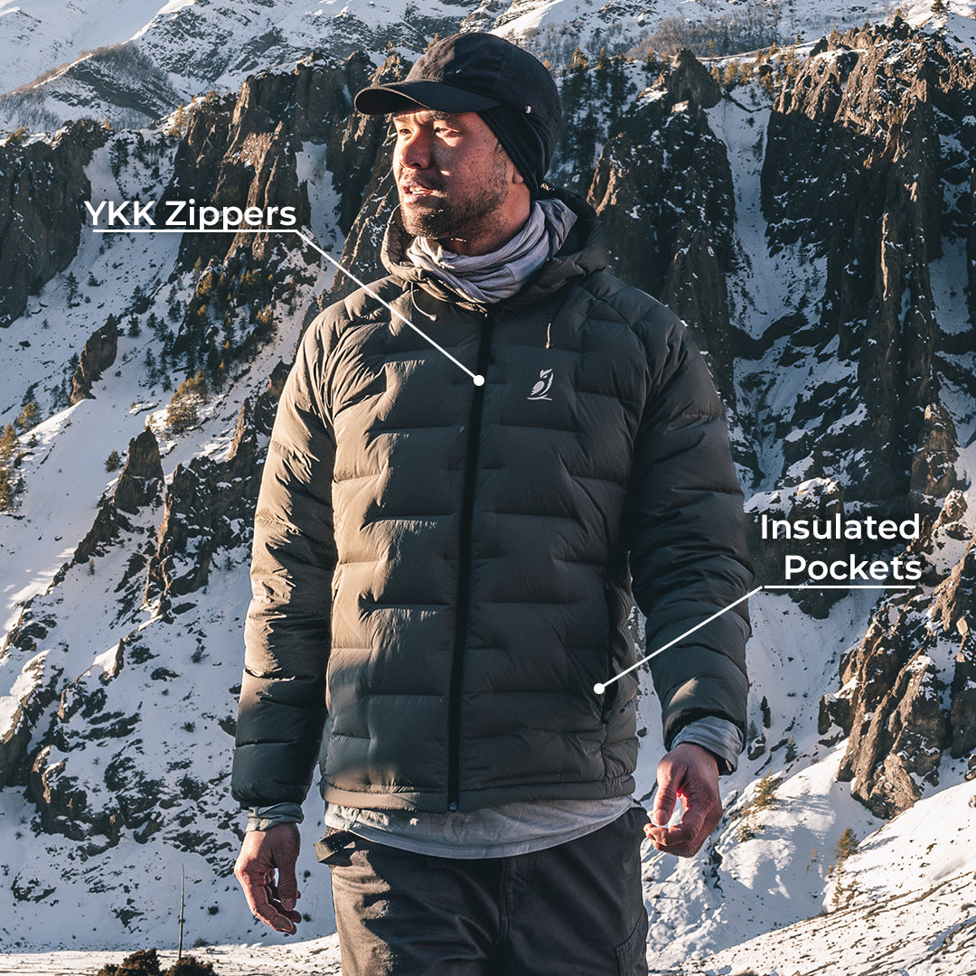 A man wearing a gray insulated puffer jacket stands in a snowy mountain landscape. White text labels point to the jacket, reading 'YKK Zippers' near the chest zipper area and 'Insulated Pockets' near the lower pocket area.